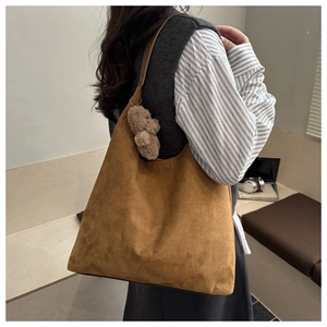 Brown faux suede tote bag with plush bear charm worn by a model in a cozy indoor setting