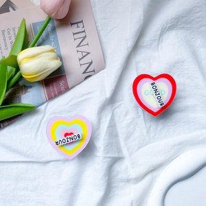 Close-up of heart-shaped acrylic phone grip with Bonjour text on white textured cloth