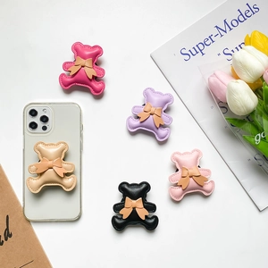 Cute 3D bow bear retractable phone socket grip stand in multiple colors shown on a desk