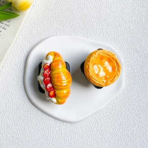 Top view of creative croissant and egg tart phone grips on a minimalist white ceramic plate