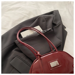 Detail view of dark red patent leather handbag with silver logo plate on grey blazer