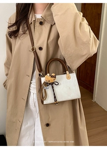 Sac bandoulière blanc avec poignée marron et pendentif peluche porté sur un trench-coat beige