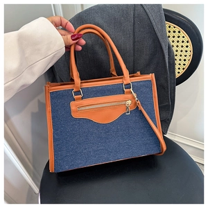 Close-up of contrast color denim tote bag featuring brown leather trim and front zip pocket