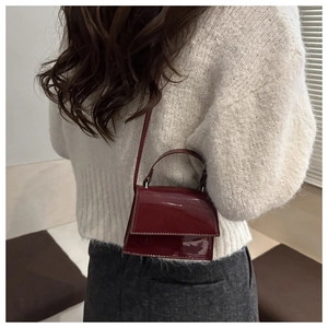 Model wearing a burgundy patent leather crossbody bag with a glossy trapezoid flap design