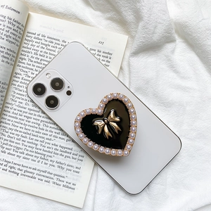 Elegant heart-shaped phone grip with gold bow and pearl inlay on a white smartphone