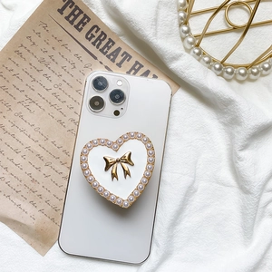 Detail of heart-shaped phone grip with gold bow and pearl trim on a white smartphone