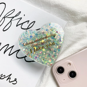 Close-up of glitter heart phone grip with gold lettering on a soft fabric background