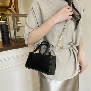 Minimalist black patent leather tote bag with gold chain strap on model in cafe