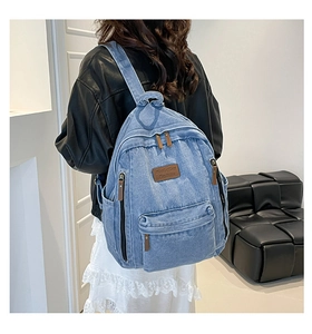 Detail view of light blue denim backpack showing multi-zipper pockets and brown leather tags