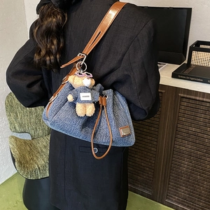 Blue denim bucket bag with brown leather strap and teddy bear charm shown in a modern interior