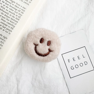 Close-up of a plush beige smiley face phone grip on a white aesthetic background