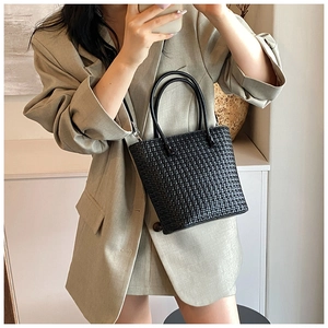 Detail view of a black woven trapezoid handbag with dual handles and slim shoulder strap