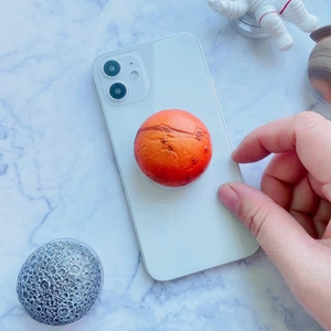 Orange planet-shaped expanding phone grip stand with textured surface on a white smartphone