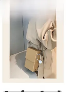 Mini beige square box crossbody bag with thin metal chain strap shown on a beige trench coat