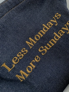 Detailed close-up of yellow 'Less Mondays More Sundays' embroidery on blue denim fabric