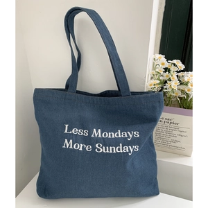 Detail of blue denim tote bag featuring white embroidered Less Mondays More Sundays text