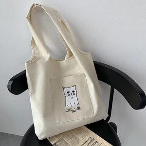Chic beige canvas tote bag with cute cat illustration and front pocket on a modern chair