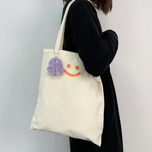 Detail of canvas tote bag featuring an orange smiley face and purple plush charm accessory
