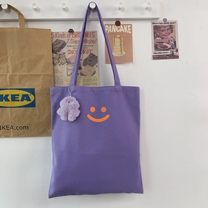 Detail of purple canvas tote bag featuring orange smiley face and plush monster charm