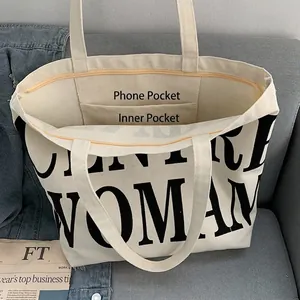 Cream canvas tote bag with black WOMAN print and zipper closure on a grey sofa