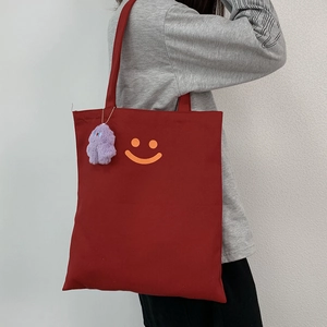 Red canvas tote bag with orange smiley face and purple plush monster charm on shoulder