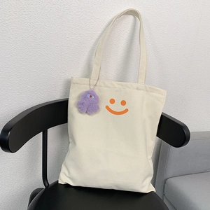 Detail of off-white canvas tote bag featuring an orange smiley face and purple plush charm