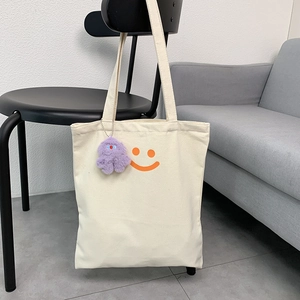 Detail of off-white canvas tote bag with orange smiley face and purple plush charm
