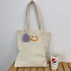 Close-up of a cream canvas tote with orange smiley face and purple plush monster charm