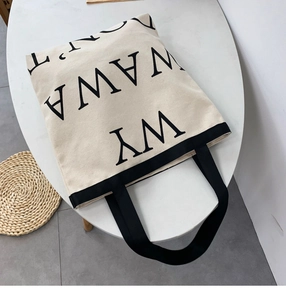 Detailed flat lay of white canvas tote bag with bold black letter print and woven handles