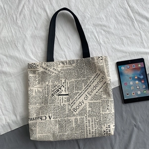Close-up of newspaper print canvas tote bag with black straps and tablet for size reference