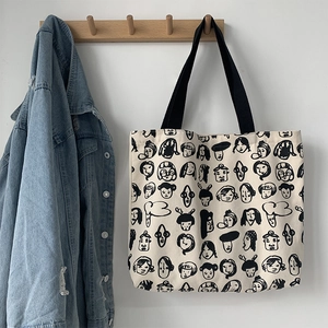 Large beige canvas tote bag with black cartoon face prints hanging on a wooden hook