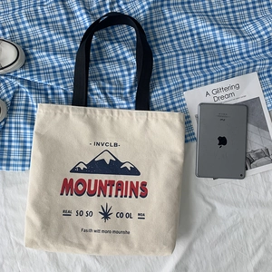 Flat lay of off-white canvas tote bag with black straps and mountain print on grid background
