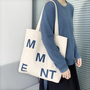 Minimalist canvas tote bag with large blue MMENT letters worn by a model in a cozy room