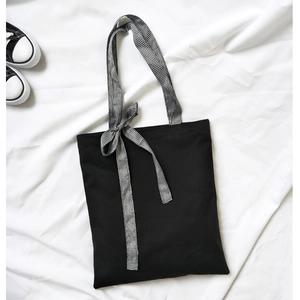 Flat lay of a black canvas tote bag with a checkered bow strap on white linen fabric