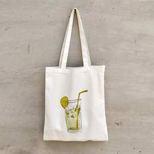 Minimalist white canvas tote bag with lemon drink illustration hanging on a neutral wall