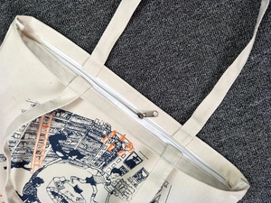 Close-up of a cream canvas tote bag showing silver zipper closure and colorful illustration