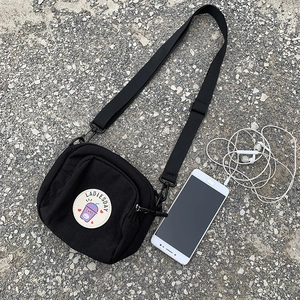 Detalle de un mini bolso bandolera de lona negra con parche y correa ajustable nylon
