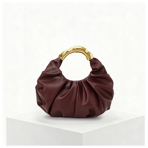 Detail of burgundy pleated dumpling bag featuring a curved gold metal handle on a white stand