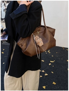 Detail of dark brown leather tote bag with plush bear charm worn by a model in outdoor setting