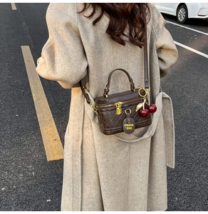 Détail du sac boîte matelassé marron foncé avec breloque cerise porté en bandoulière par le mannequin