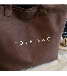 Close-up of a dark coffee tote bag featuring white TOTE BAG text on suede texture