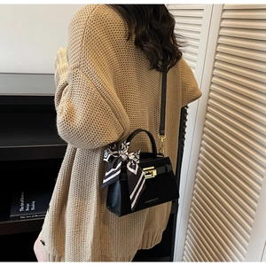 Detail of black leather trapezoid bag with gold clasp and silk scarf on a beige cardigan