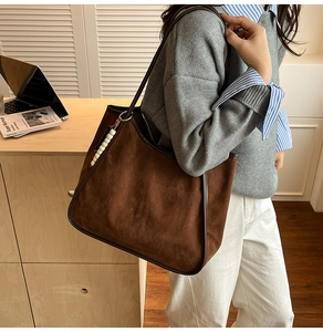 Brown suede tote bag with leather trim and beaded charm worn by a model in warm light