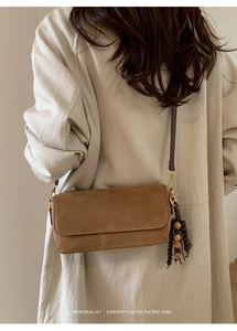 Brown suede crossbody bag with decorative wooden bead tassel shown on a beige trench coat