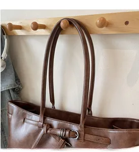 Detail of a brown leather tote bag with metallic buckle and long straps on a wooden hook