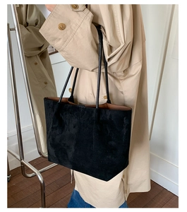 Detail of a black suede tote bag with slim shoulder straps worn by a model in a minimal room