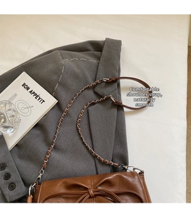 Detalle de un bolso marrón con cadena y lazo decorativo sobre una chaqueta gris