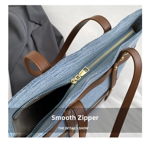 Close-up of golden metal zipper and brown leather trim on a blue denim tote bag