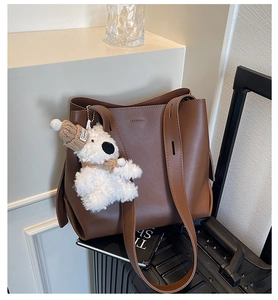 Detail of a brown leather tote bag featuring a cute teddy bear plush charm and wide strap