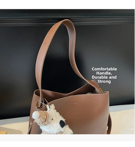 Close-up of brown leather bucket bag featuring a wide shoulder strap and bear charm